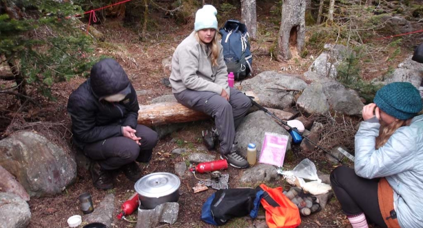 A group of people sit at a campsite in a wooded area and prepare food